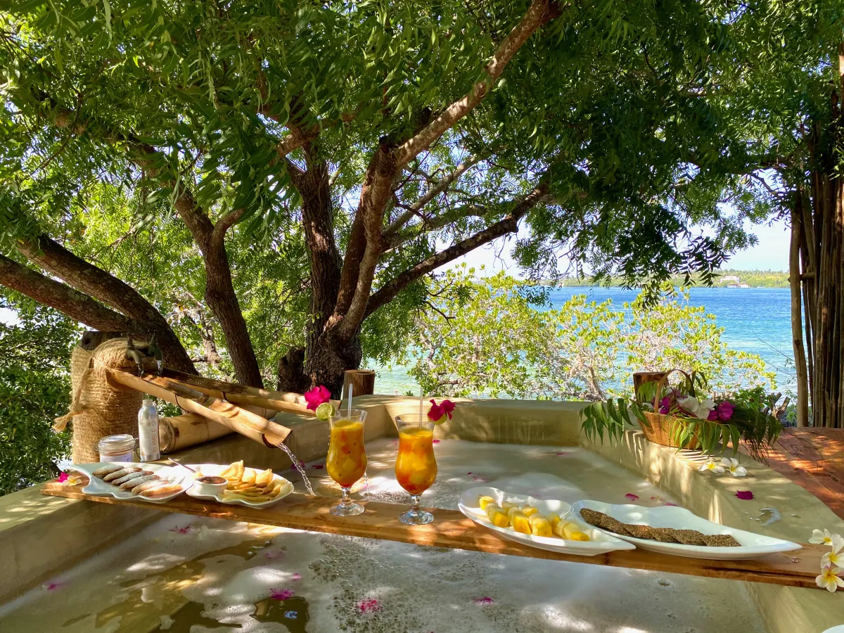 A private outdoor bath scattered with flower petals, breakfast and juices served on a tray beneath a tree by the water