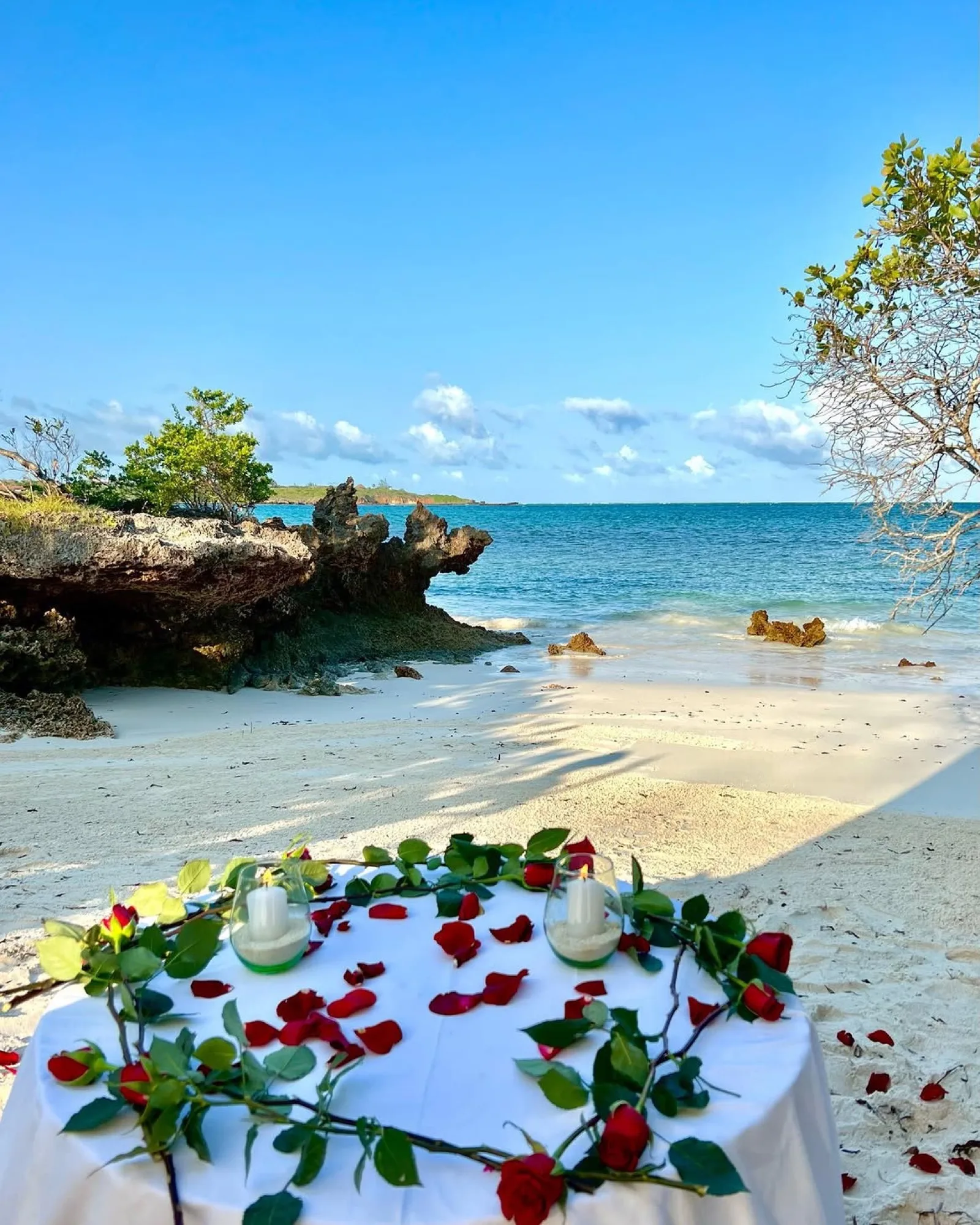 A table for two dressed with red roses and candles on the white sand at the water’s edge